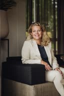 Tanja zit glimlachend in een fauteuil bij het raam, gekleed in een licht broekpak.