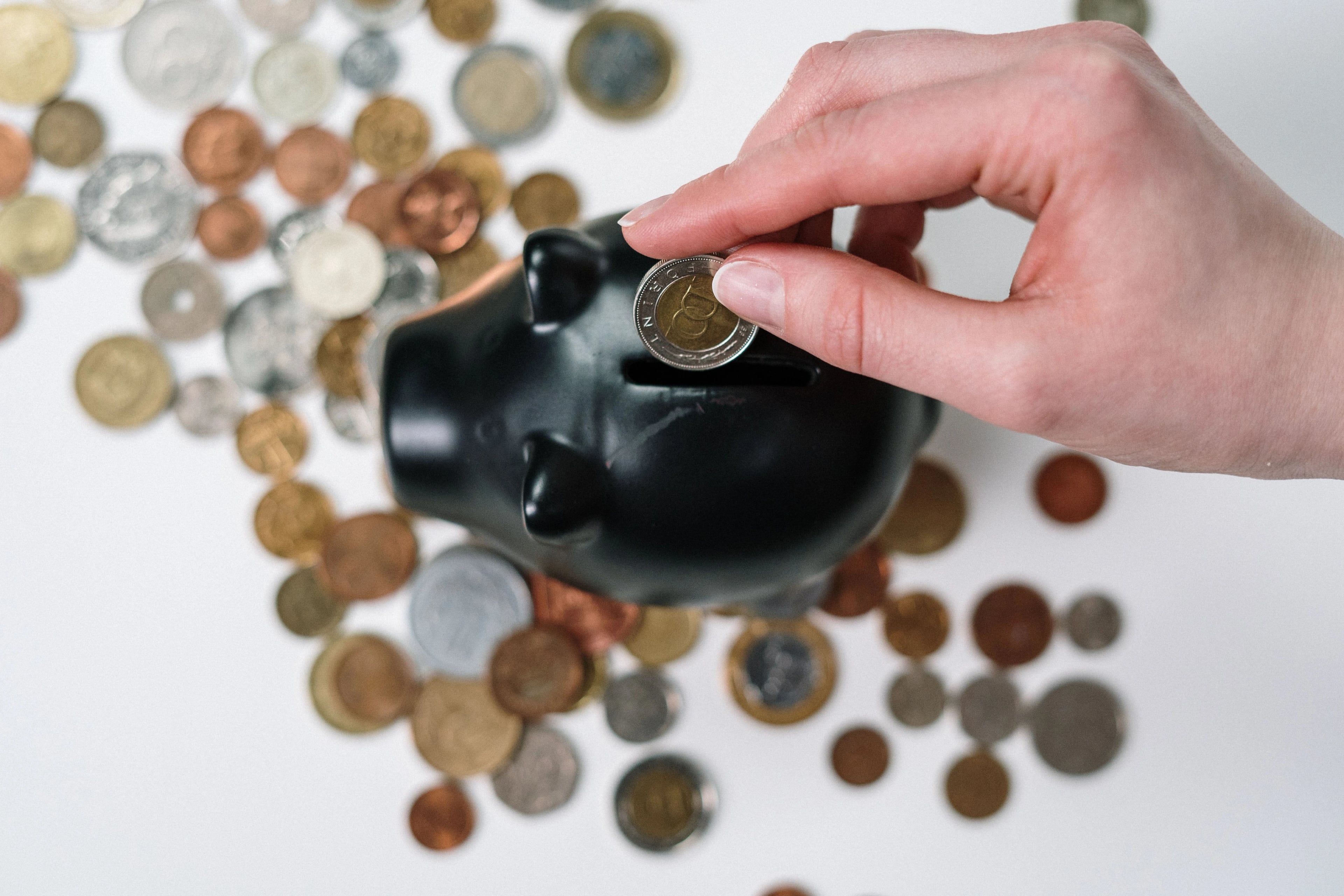 Hand puts a coin into a black piggy bank, surrounded by coins on a white surface.